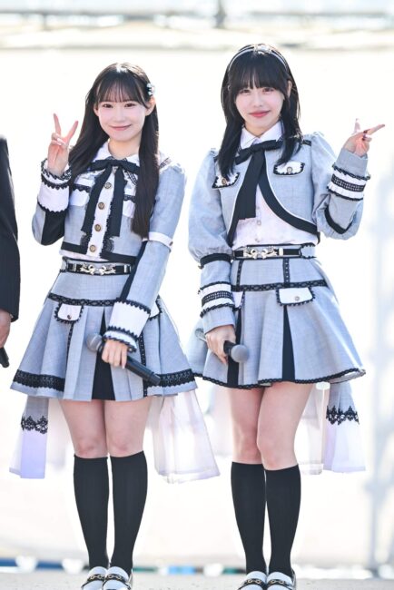 SKE48の佐藤佳穂と大村杏が、屋外ステージでグレーと白のアイドル衣装を着てポーズを取っている。佐藤佳穂はピースサイン、大村杏は指でピースサインをしている。