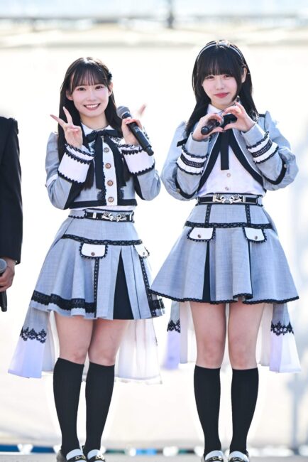 SKE48の佐藤佳穂と大村杏が、屋外ステージでグレーと白のアイドル衣装を着てポーズを取っている。佐藤佳穂はピースサイン、大村杏は指でハートマークを作っている。