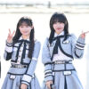 SKE48の佐藤佳穂と大村杏が、屋外ステージでグレーと白のアイドル衣装を着てポーズを取っている。佐藤佳穂はピースサイン、大村杏は指でハートマークを作っている。